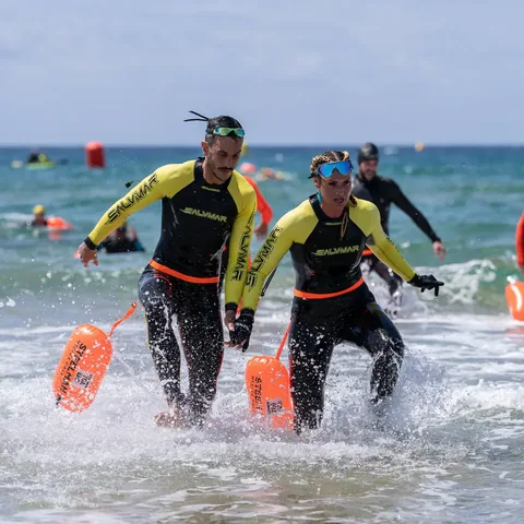 Two athletes in wetsuits running out of the sea with tow buoys.